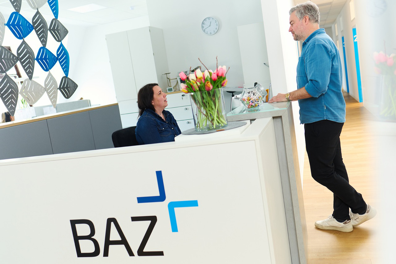 Mann in blauem Hemd steht am Empfang der Arztpraxis und spricht mit der medizinischen Fachangestellten